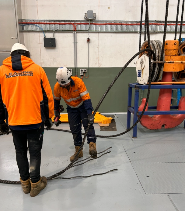Technicians inspecting and handling crane wire rope near hoist drum in industrial workshop setting